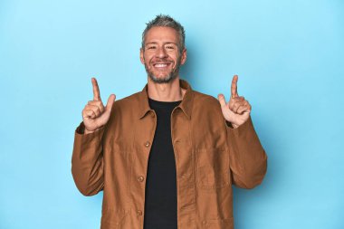 Mavi arka plandaki orta yaşlı beyaz adam iki ön parmağı da boş bir alan gösteriyor..