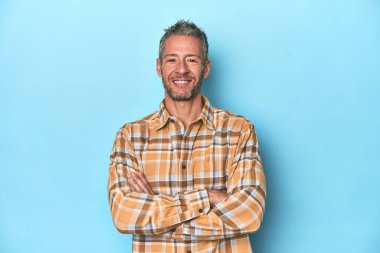 Mavi arka planda oturan orta yaşlı beyaz bir adam kendinden emin, kararlılıkla kollarını kavuşturuyor..