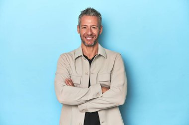 Mavi arka planda oturan orta yaşlı beyaz bir adam kendinden emin, kararlılıkla kollarını kavuşturuyor..