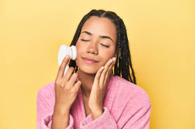 Pembe cüppeli genç Endonezyalı sarı stüdyo arka planında yüz temizleyicisi kullanıyor.