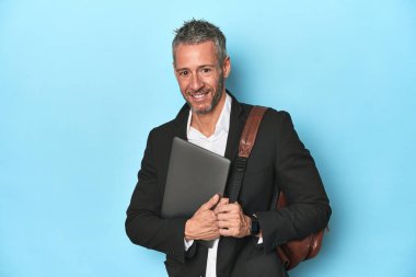 Sırt çantalı bir iş adamı dizüstü bilgisayarını mavi arkaplanda tutuyor.