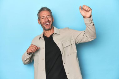 Mavi arka plandaki orta yaşlı beyaz adam özel bir günü kutluyor, atlıyor ve enerjiyle kollarını kaldırıyor..