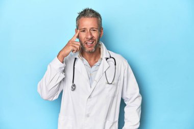 Mavi arka planda orta yaşlı beyaz bir doktor işaret parmağıyla bir hayal kırıklığı gösteriyor..
