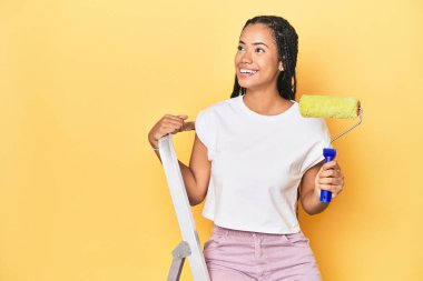 Endonezyalı kadın merdivenli ve sarı arka planda boya silindirli.