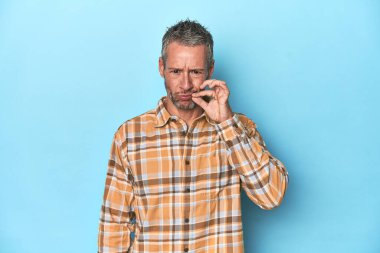 Orta yaşlı beyaz bir adam mavi arka planda parmakları dudaklarında bir sır saklıyor..