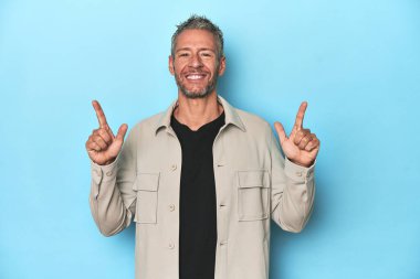 Mavi arka plandaki orta yaşlı beyaz adam iki ön parmağı da boş bir alan gösteriyor..