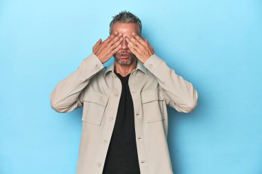 Mavi arka planda orta yaşlı beyaz bir adam elleriyle gözlerini kapatmaktan korkuyor..