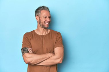 Mavi arka planda oturan orta yaşlı beyaz adam kollarını kavuşturarak gülümsüyor..