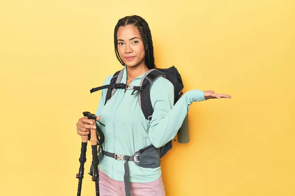 黄色のハイキングギアを持つインドネシアの女性は 手のひらにコピースペースを表示し 腰に別の手を保持 — ストック写真