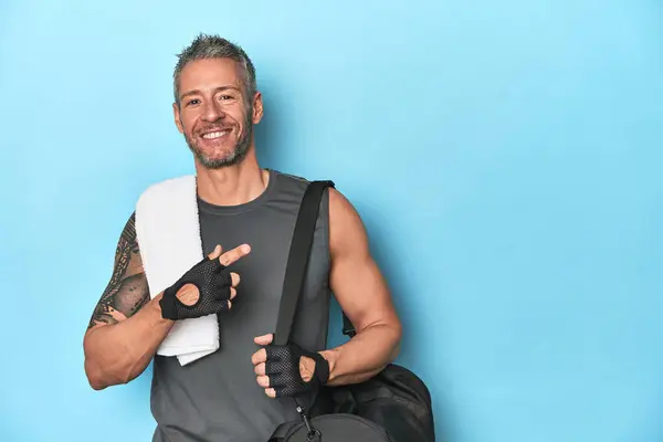 Athlete with gym backpack on blue background smiling and pointing aside, showing something at blank space.