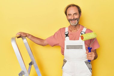 Orta yaşlı bir adam merdivenle sarı bir duvar boyuyor.