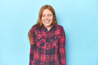 Mavi arka plandaki kızıl saçlı genç kadın gülüyor ve gözlerini kapatıyor, rahatlamış ve mutlu hissediyor..