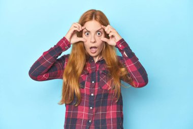 Mavi arka plandaki kızıl saçlı genç kadın bir başarı fırsatı bulmak için gözlerini açık tutuyor..