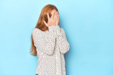 Mavi arka plandaki kızıl saçlı genç kadın korkmuş ve gergin parmaklarıyla göz kırpıyor..