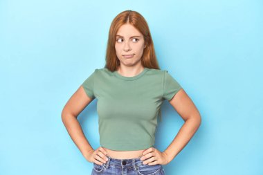 Mavi arka plandaki kızıl saçlı genç kadın şaşkın, kuşkulu ve güvensiz hissediyor..