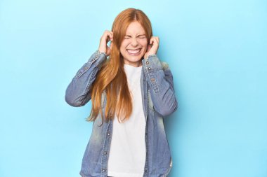 Mavi arka planda kızıl saçlı genç bir kadın kulaklarını elleriyle kapatıyor..