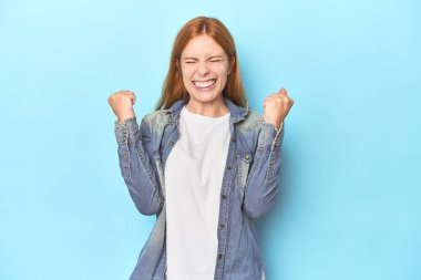 Mavi arka plandaki kızıl saçlı genç kadın tasasız ve heyecanlı bir şekilde tezahürat yapıyor. Zafer konsepti.