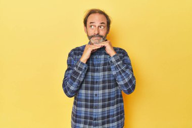 Sarı stüdyodaki orta yaşlı beyaz adam ellerini çenesinin altında tutuyor ve mutlu bir şekilde yan tarafa bakıyor..