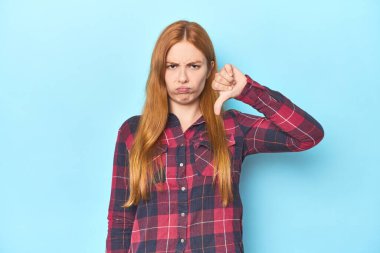 Mavi arka plandaki kızıl saçlı genç kadın baş parmağını aşağı gösteriyor, hayal kırıklığı konsepti..