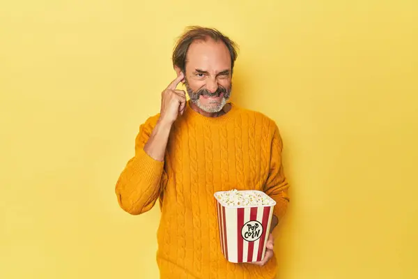 Człowiek Cieszący Się Popcorn Żółtym Studio Przykrywając Uszy Rękami — Zdjęcie stockowe