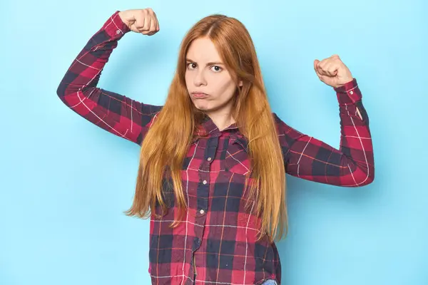 Mavi arka plandaki kızıl saçlı genç kadın güçlü bir hareket gösteriyor, kollarıyla, kadınsı gücün sembolü.