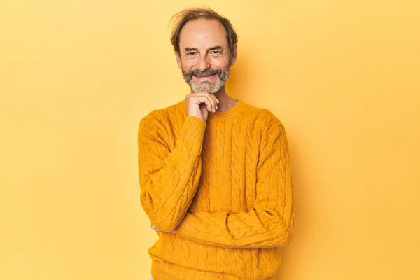 Caucasian middle-aged man in yellow studio smiling happy and confident, touching chin with hand.
