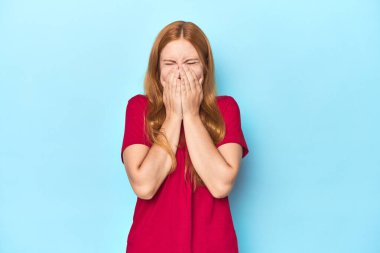Mavi arka plandaki kızıl saçlı genç kadın bir şeye gülüyor, elleriyle ağzını kapatıyor..