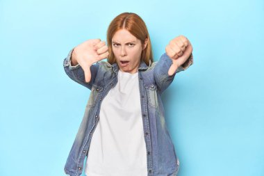 Mavi arka plandaki kızıl saçlı genç kadın baş parmağını aşağı gösteriyor ve hoşlanmadığını ifade ediyor..