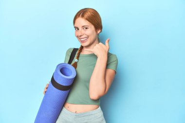 Stüdyoda yoga minderi tutan kızıl saçlı genç bir kadın cep telefonu hareketini parmaklarıyla gösteriyor..