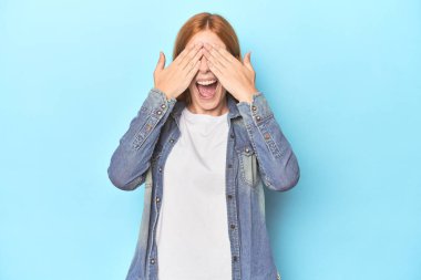 Mavi arka plandaki kızıl saçlı genç kadın elleriyle gözlerini kapatıyor. Gülümsüyor ve bir sürpriz bekliyor..