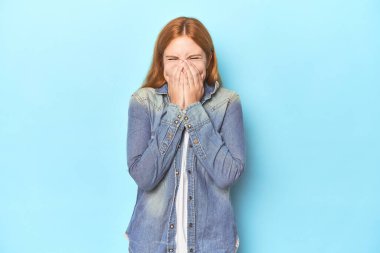 Mavi arka plandaki kızıl saçlı genç kadın bir şeye gülüyor, elleriyle ağzını kapatıyor..