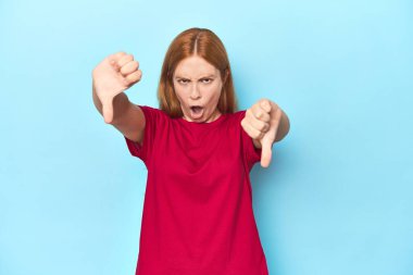 Mavi arka plandaki kızıl saçlı genç kadın baş parmağını aşağı gösteriyor ve hoşlanmadığını ifade ediyor..