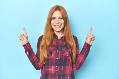 Mavi arka plandaki kızıl saçlı genç kadın iki ön parmağı da boş bir alan gösteriyor..