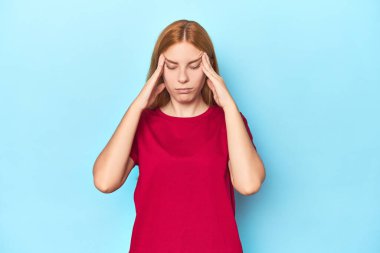 Mavi arka plandaki kızıl saçlı genç kadın şakaklara dokunuyor ve baş ağrısı çekiyor..