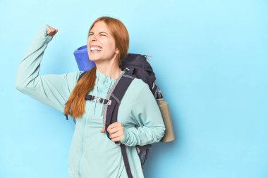 Stüdyoda sırt çantası ve paspas taşıyan kızıl saçlı gezgin zaferden sonra yumruğunu kaldırıyor, kazanan konsept.