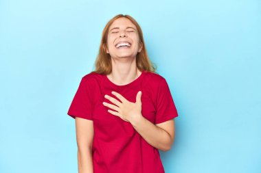 Mavi arka plandaki kızıl saçlı genç kadın elini göğsünde tutarak kahkahalar atıyor..