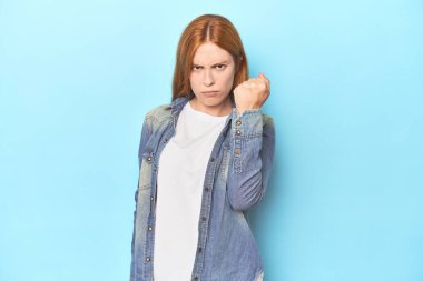 Mavi arka planda kızıl saçlı genç bir kadın kameraya yumruğunu gösteriyor, agresif bir yüz ifadesi..