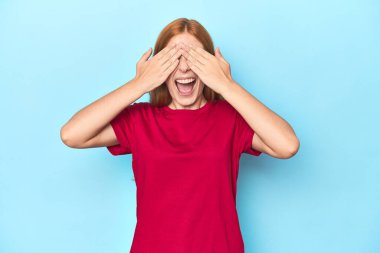 Mavi arka plandaki kızıl saçlı genç kadın elleriyle gözlerini kapatıyor. Gülümsüyor ve bir sürpriz bekliyor..