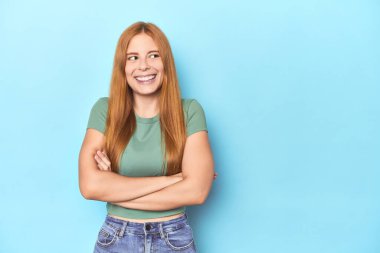 Mavi arka plandaki kızıl saçlı genç kadın kollarını kavuşturarak gülümsüyor..