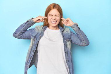 Mavi arka planda, kulaklarını parmaklarıyla kapatan kızıl saçlı genç bir kadın gürültülü bir ortam yüzünden stresli ve çaresiz..