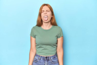 Mavi arka planda kızıl saçlı genç bir kadın komik ve arkadaş canlısı bir dil uzatıyor..
