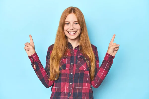 蓝色背景的红头发年轻女子用两个前指竖起来表示空白 — 图库照片