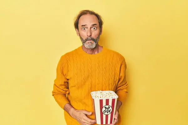 Człowiek Cieszący Się Popcorn Żółtym Studio Wzrusza Ramionami Otwarte Oczy — Zdjęcie stockowe