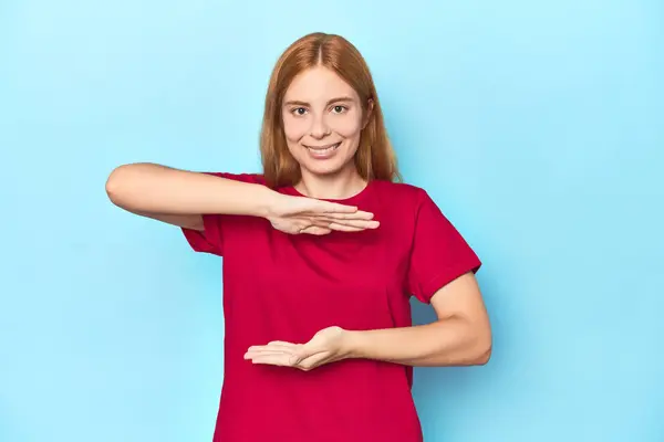 両手で何かを持っている青い背景の赤毛の若い女性 製品プレゼンテーション — ストック写真
