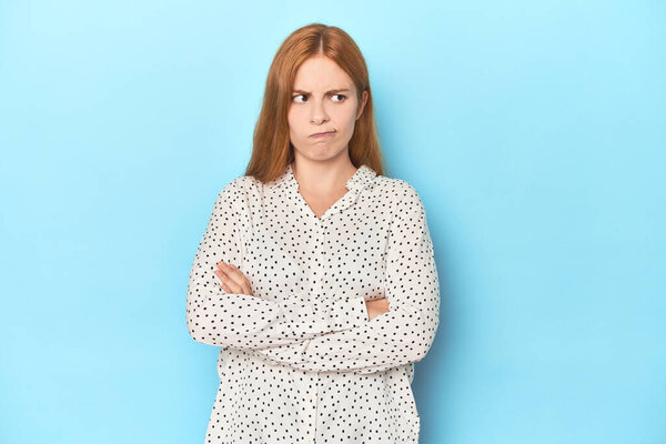 Рыжая молодая женщина на синем фоне хмурящаяся в неудовольствие, держит руки сложенными.