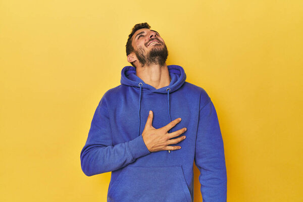 Young Hispanic man on yellow background laughs out loudly keeping hand on chest.