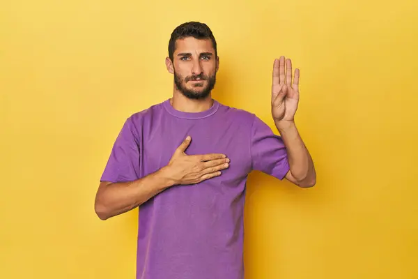 年轻的有黄色背景的西班牙裔男子宣誓 把手放在胸前 — 图库照片