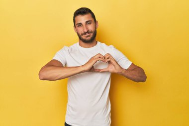 Sarı arka planda gülümseyen ve elleriyle kalp şeklini gösteren genç bir İspanyol..