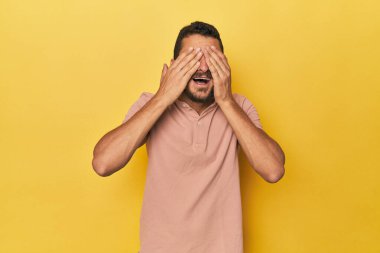 Sarı arka plandaki genç İspanyol adam elleriyle gözlerini kapatıyor. Gülümsüyor ve bir sürpriz bekliyor..