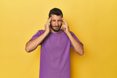 Sarı arka plandaki genç İspanyol adam şakaklara dokunuyor ve baş ağrısı çekiyor..